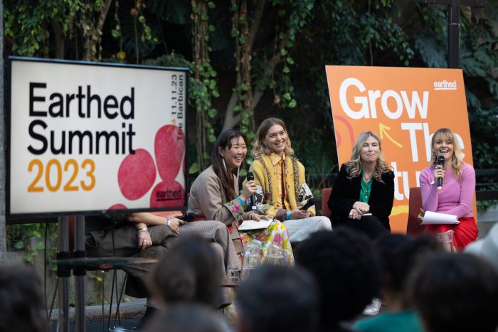 Earthed Summit 2024 panel, 4 panelists sit on a stage, surrounded by lush green setting of The Barbican in London. They hold microphones and laugh.